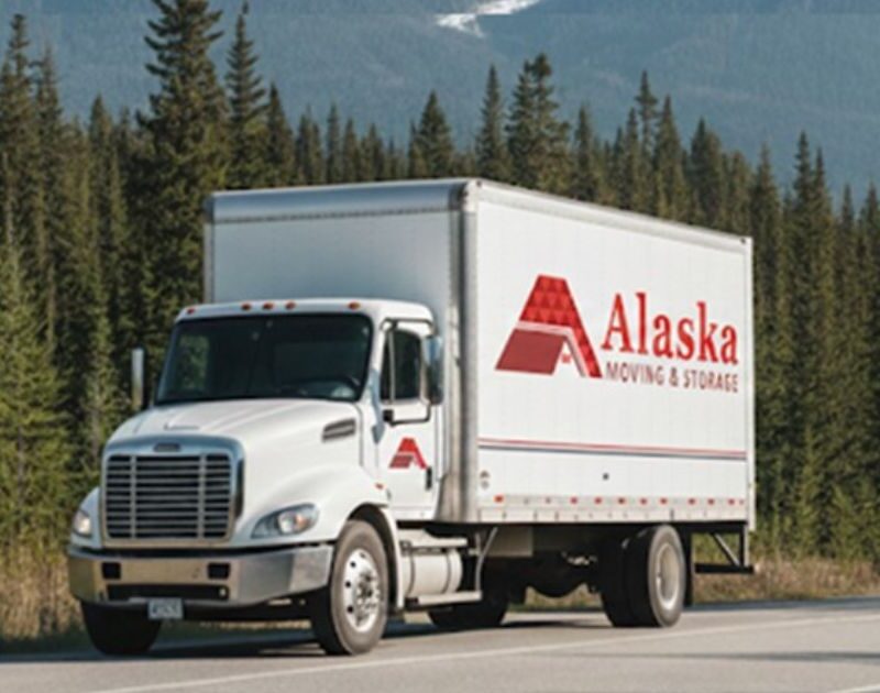 Alternative Text Alaska Moving & Storage truck driving on a road surrounded by evergreen pine trees and snowy mountains